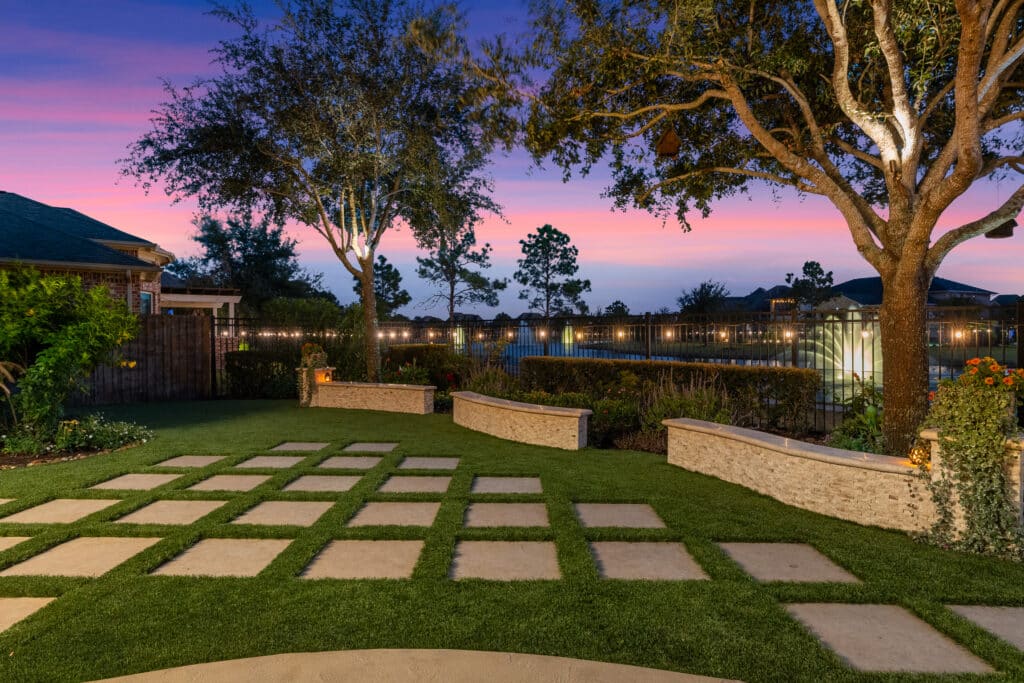 Artificial turf lawn with stone pavers in a landscaped backyard at dusk